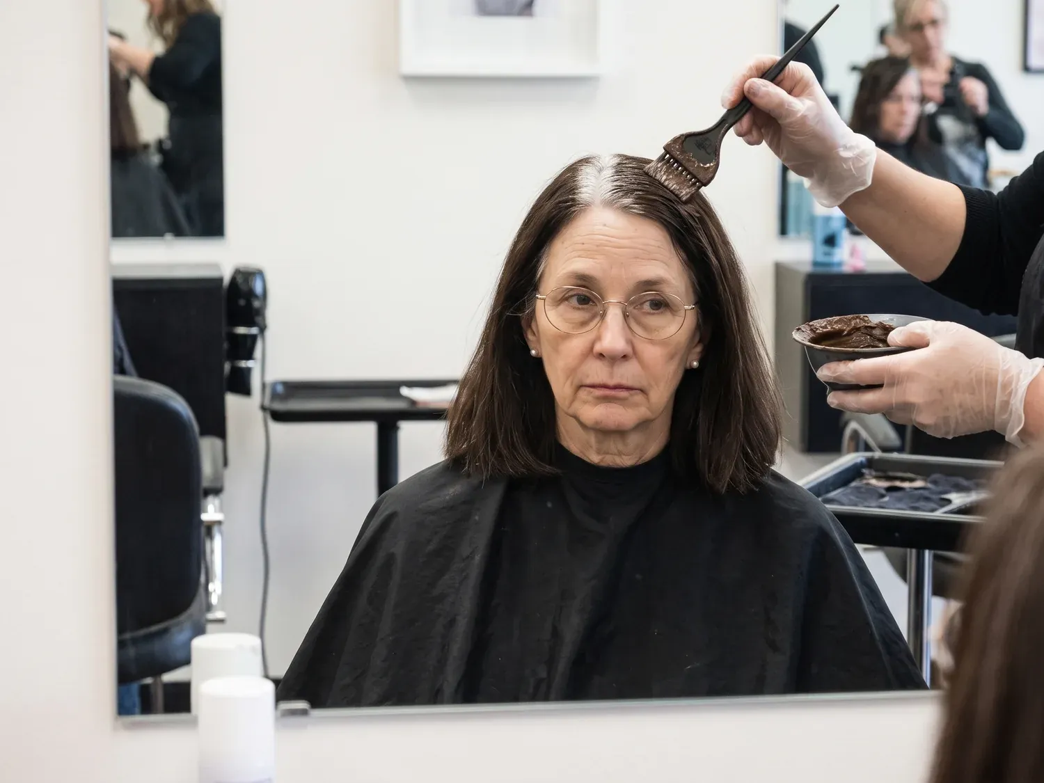 A woman in her sixties in a salon chair under a black cape while a colorist brushes dark color paste onto her roots