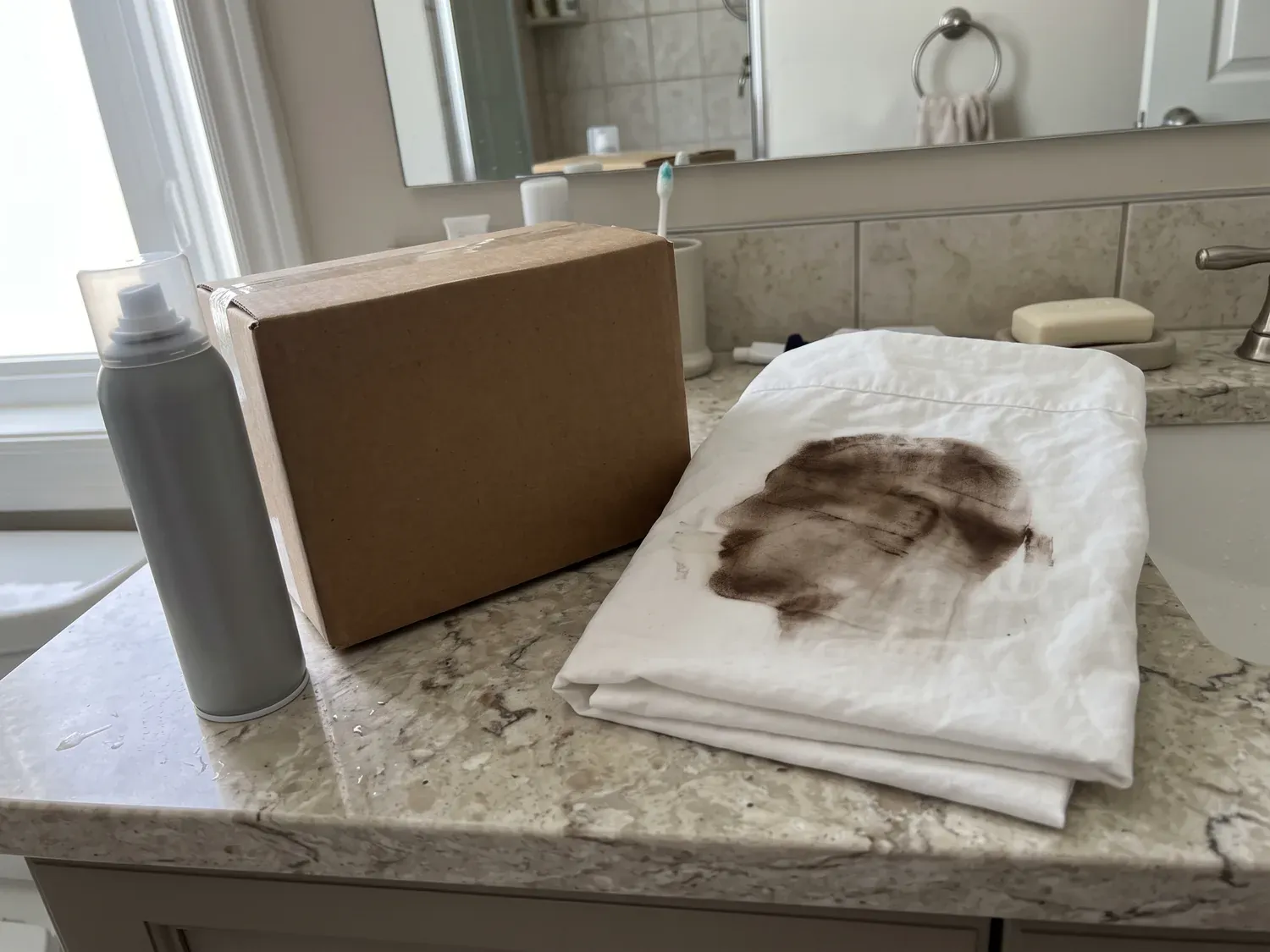 A bathroom counter with a plain box, an unlabeled root spray can and a pillowcase with a brown smudge