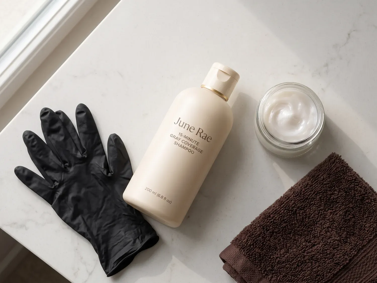 Flat lay on a bathroom counter: the June Rae bottle, black disposable gloves, a small jar of rich cream and a folded dark brown towel